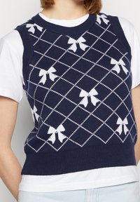 Marineblauer Strickpullover mit weißen Schleifenmustern, Rautengitterdesign, geripptem Kragen und Saum, getragen über einem weißen T-Shirt.