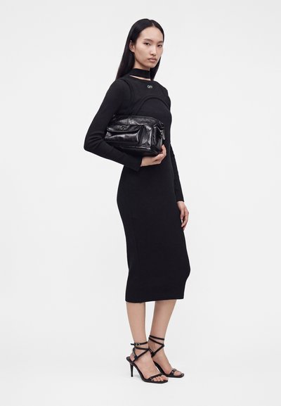 Femme en robe noire à manches longues et talons à brides tenant un sac à main en cuir noir sur un fond clair.