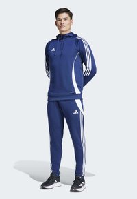Jūrinis sportinis komplektas su kontrastingomis baltomis juostelėmis, užtraukiama gobtuvu viršutine dalimi ir siaurėjančiomis kelnėmis. Matinė medžiaga su įspaustu logotipo elementu.