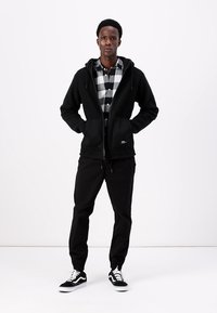 Veste en polaire noire zippée avec deux poches avant, superposée à une chemise à carreaux noir et blanc, associée à un pantalon noir ajusté et des baskets noires.