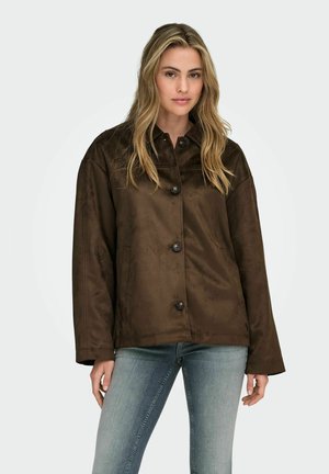 Naine, kellel on pikad blondid juuksed, seljas kinnitatud pruun sametjakk ja helesinised teksad, sirge heleda taustaga.