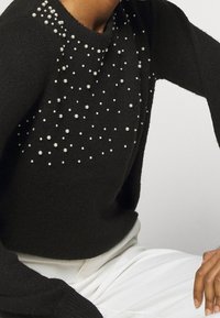 Svart tröja med utspridda små vita pärlor, parat med vita byxor, visande torso och höger hand som vilar på knäet.