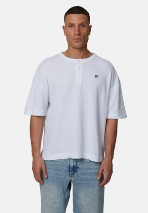 Maglietta henley bianca a maniche corte, realizzata in tessuto a coste; presenta una scollatura rotonda, una patta con tre bottoni e un piccolo dettaglio con il logo.