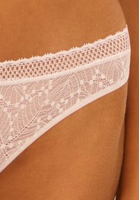 String en dentelle rose pâle avec des motifs floraux complexes, doté d'une taille en dentelle à bords festonnés. Finition lisse et détails délicats.