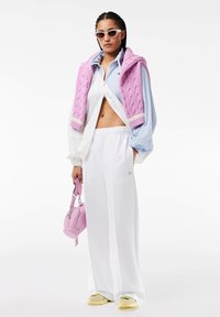 Pantaloni albi, bluză cu fermoar de culoare albastru deschis, pulover tricotat roz pe umeri, geantă pastel roz, adidași galbeni și ochelari de soare albi.