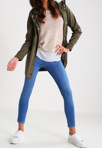 Parka vert olive, pull beige texturé, jean skinny bleu et baskets blanches ; tenue décontractée avec un design superposé.