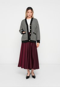 Woman wearing black and white houndstooth cardigan with gold buttons, white top, burgundy satin midi skirt, and black pointed slingback shoes.