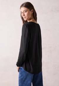Blusa nera a maniche lunghe con tessuto testurizzato e scollatura rotonda, caratterizzata da una vestibilità rilassata e una leggera drappeggiatura sul retro. Abbinata a jeans blu.