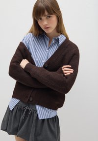 Cárdigan marrón de punto con escote en V y cierre de botones, superpuesto a una camisa de rayas azul claro y blanca, combinado con una falda gris.