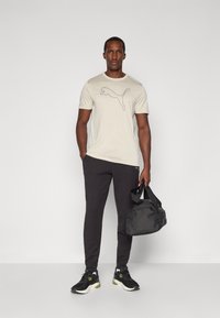 Lichtbeige t-shirt met zwart Puma-logo, grijze tapered broek en zwarte sneakers met groene accenten, plus een zwarte sporttas.