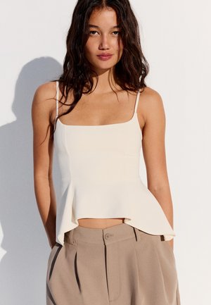 Jeune femme aux cheveux foncés lâchés porte un haut blanc sans manches avec fines bretelles et un pantalon beige taille haute sur un fond uni.