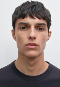 Giovane uomo con capelli corti e scuri e un'espressione neutra, che indossa una maglietta nera a collo rotondo su uno sfondo grigio chiaro.