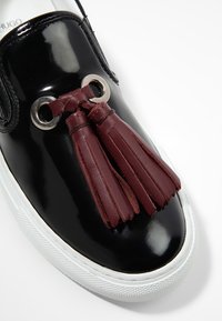 Baskets en cuir verni noir avec un détail pompon en cuir bordeaux et des accessoires argentés, dotées d'une semelle en caoutchouc blanche texturée.