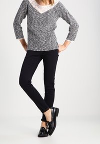 Svart och vit mönstrad långärmad topp med spetsnedre, kombinerad med svarta slim-fit byxor och glänsande svarta skor.