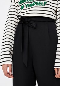 Pantalon noir taille haute avec un devant plat et une ceinture nœud papillon. Associé à un haut à manches longues crème et noir à rayures avec un texte vert.