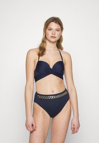 Conjunto de biquíni azul-marinho com um top de design torcido e parte de baixo de cintura alta adornada com um detalhe de corrente dourada na cintura. Textura suave.