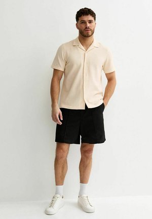 Uomo in piedi con una mano in tasca, che indossa una camicia beige a maniche corte con bottoni, pantaloncini neri, calzini bianchi e scarpe da ginnastica bianche su sfondo bianco.