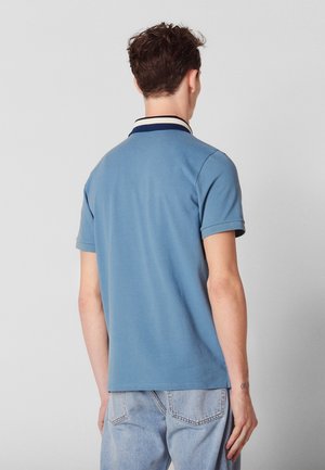 Hombre joven de espaldas que lleva una camiseta polo azul de manga corta con cuello a rayas y vaqueros azul claro.