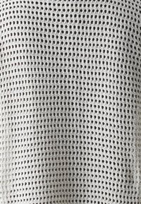 Lehká, otevřená pletenina v bílé barvě s mřížkovým vzorem. Textura je vzdušná a umožňuje viditelnost skrze síťovitou konstrukci.