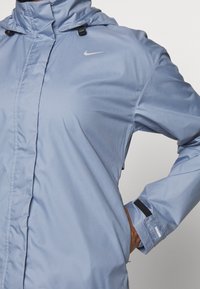 Persona con chaqueta con capucha Nike de color azul claro, con cremallera frontal y correa ajustable en la muñeca, con la mano en el bolsillo contra un fondo sencillo.