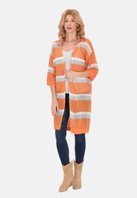 Hosszú narancssárga kötött kardigán fehér csíkokkal, laza fazonnal és háromnegyedes ujjakkal, sötét farmerral és világosbarna bokacsizmával párosítva.