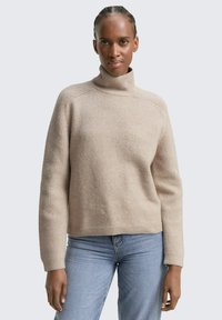 Beżowy dzianinowy sweter z wysokim kołnierzem, prążkowanymi mankietami i dolnym wykończeniem oraz miękką teksturą. Połączony z jasnoniebieskimi dżinsami w luźnym kroju.