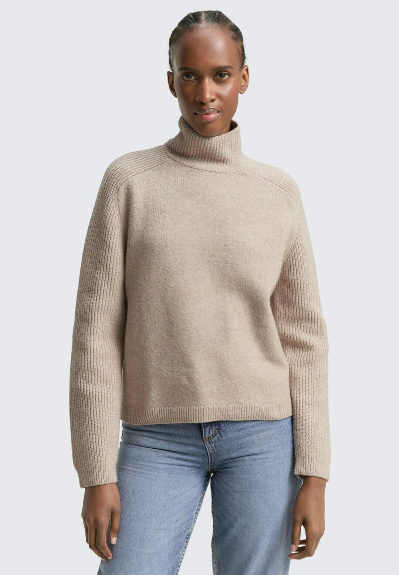 Beżowy dzianinowy sweter z wysokim kołnierzem, prążkowanymi mankietami i dolnym wykończeniem oraz miękką teksturą. Połączony z jasnoniebieskimi dżinsami w luźnym kroju.