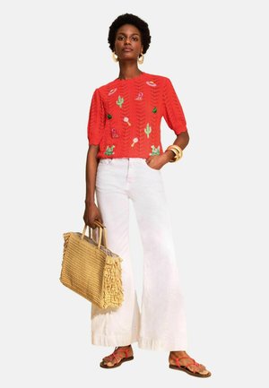 Woman wearing a red short-sleeve sweater with embroidered cacti and objects, white wide-leg pants, red sandals, gold hoop earrings, and holding a woven bag.