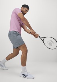 Pólder de manga corta rosa claro, pantalones cortos grises, zapatos deportivos blancos con detalles azules, sosteniendo una raqueta de tenis, preparado para golpear una pelota.
