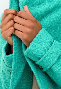 Cappotto turchese con texture e dettagli a bottoni. Il tessuto appare morbido con una finitura pelosa. Mani afferrano il tessuto, mettendo in risalto il calore e il comfort.