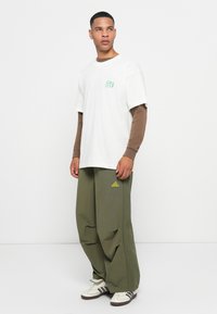 Witte katoenen T-shirt, groene grafische logo; bruine lange mouwen eronder. Olijfgroene broek met zijstrepen, gele merklogo. Sneakers met strepen.
