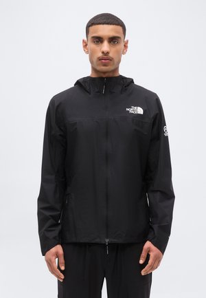 Mann med svart The North Face jakke med glidelås, hette og logo på brystet og høyre overarm, står foran en ensfarget bakgrunn.