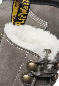 Stivale in pelle grigia con rivestimento in pelliccia bianca, lacci grigi e occhielli in metallo. Etichetta in tessuto giallo con testo nero attaccata alla parte superiore dello stivale.