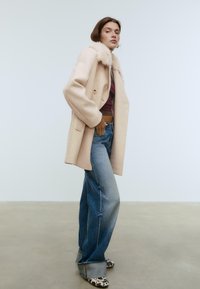 Beige wollen jas met een nepkraag van bont, gecombineerd met hoge blauwe jeans en gemustreerde hakken. Eenvoudig en gestructureerd ontwerp.