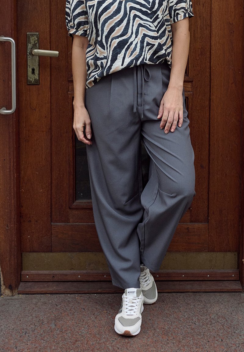 Pantalon gris à jambes larges en tissu doux, doté d'une taille à cordon et de poches latérales, associé à des baskets blanches et grises.