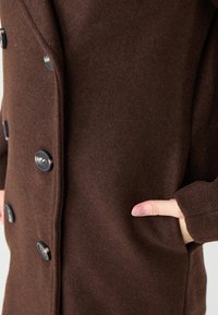 Brauner Wollmantel mit großen schwarzen Knöpfen, strukturiertem Finish, seitlichen Taschen und einem klassischen Design mit einer leicht lockeren Passform.