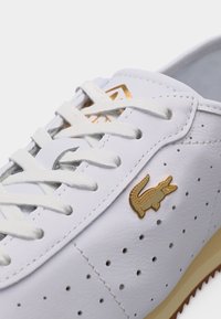 Λευκό δερμάτινο αθλητικό παπούτσι με λευκά κορδόνια, διάτρητη πλευρά, μπεζ σόλα και χρυσό λογότυπο κροκόδειλου στην πλευρική επιφάνεια.