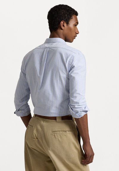 Ανοιχτό μπλε πουκάμισο με ρίγες και μακριά ρολάρισμένα μανίκια, κατασκευασμένο από βαμβάκι, συνδυασμένο με μπεζ παντελόνι που διαθέτει τσέπες και ζώνη.