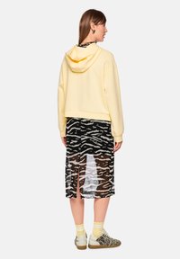 Gelber, gekürzter Hoodie mit Kordelzug an der Kapuze, kombiniert mit einem schwarzen, transparenten Rock mit weißem Zebramuster. Weiße Sneakers vervollständigen den Look.