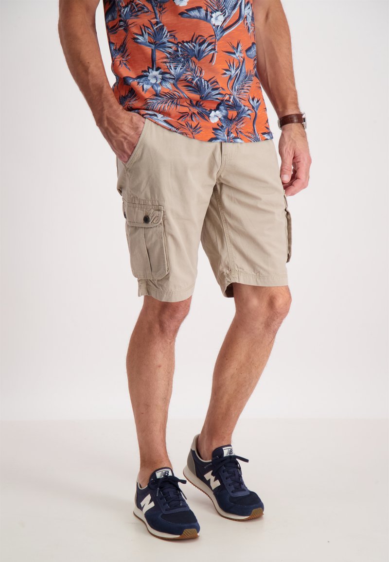 Pantaloni cargo beige con tasche laterali e una vestibilità rilassata, abbinati a una camicia floreale con una vivace fantasia arancione e blu e sneakers blu navy.