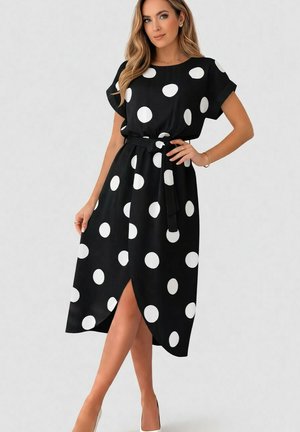 Femme portant une robe midi noire à gros pois blancs, manches courtes, taille ceinturée et fente devant, debout, main sur la hanche.