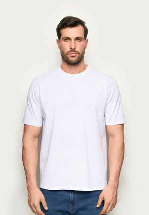 Uomo con capelli castani corti e barba, indossa una t-shirt bianca a maniche corte e jeans blu, in piedi davanti a uno sfondo chiaro.