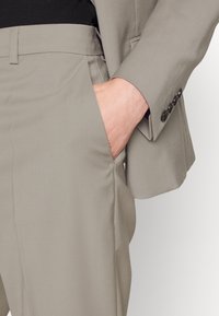 Person klädd i beige kostym med handen avslappnat vilande i byxfickan, vilket visar kostymjackans ärm med fyra svarta knappar.