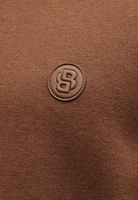 Okrogla rjava usnjena etiketa z vtisnjenim dvojnim črko B na svetlo rjavi pleteni tkanini v ozadju.