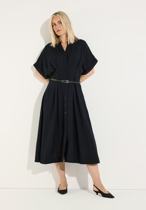 Blonde Frau mit glattem Haar, die ein schwarzes Midikleid mit Gürtel und schwarze Slingback-Schuhe mit niedrigem Absatz trägt, steht vor einer schlichten weißen Wand.