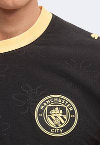 Κοντινή λήψη μαύρου αθλητικού μπλουζακιού Manchester City με ανοιχτό κίτρινο γιακά και διακριτικά μοτίβα μελισσών στο ύφασμα.