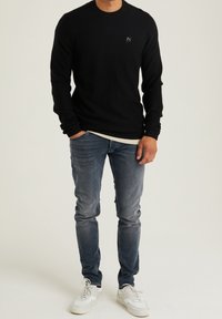 Maglione nero lavorato a maglia a maniche lunghe con scollatura rotonda e un piccolo logo, abbinato a jeans blu sbiaditi e sneakers bianche.