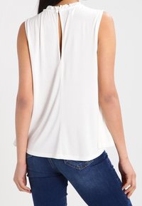 Valkoinen hihaton paita, jossa on röyhelökaulus, avattu selkäosa ja sileä pinta. Yhdistetty tummansiniset farkut.