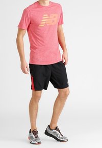 Red athletic t-shirt with yellow logo, black shorts with red side panel, and grey sneakers with orange accents.