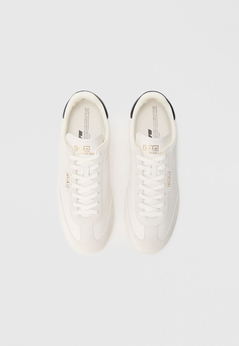 Le sneakers bianche presentano una tomaia in pelle liscia, chiusura con lacci e un accento nero sul tallone. Dettaglio del logo dorato vicino alla linguetta.